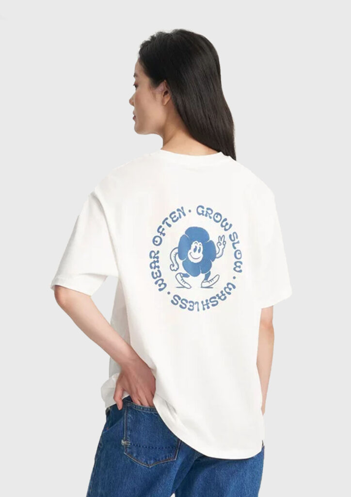 A woman models a white t-shirt containing an illustration on the reverse. The illustration is a cartoon indigo flower, walking and waving. There is typography around it, in a circle.