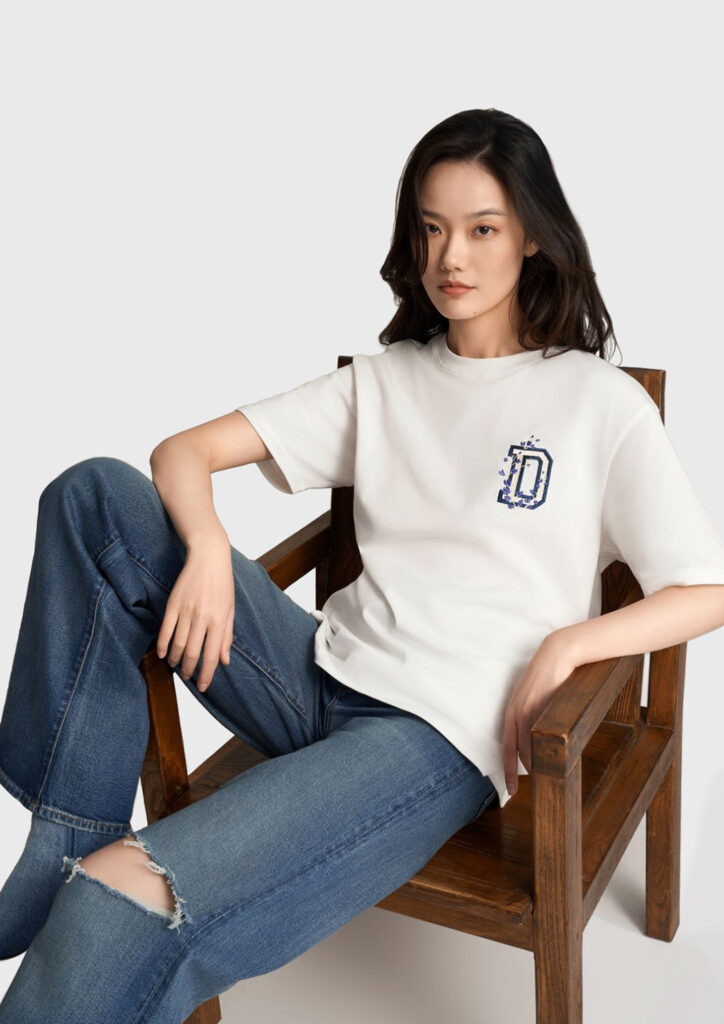 A photo of a beautiful model sat in a chair. She is wearing a white t-shirt and blue jeans. The shirt has a small image of a letter D, decorated in indigo flowers.