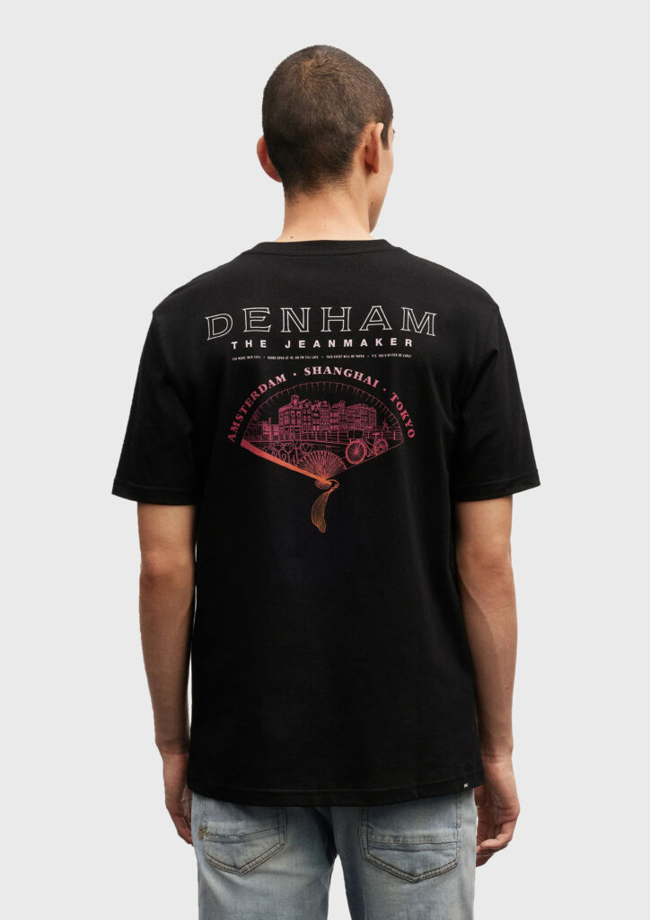 A model wearing a Denham shirt, featuring a design of a Japanese folding fan, containing a landscape scene of Amsterdam. The illustration has a gradient colour effect on it, and it has typography above it.