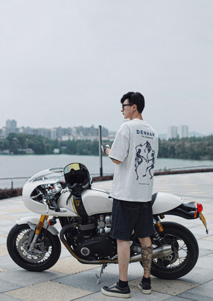 A man modelling a boxy fit Denham t-shirt. The shirt is white with a design of three, blue skeletons dancing together on the back. The male is stood next to a motorcycle with a cityscape background.